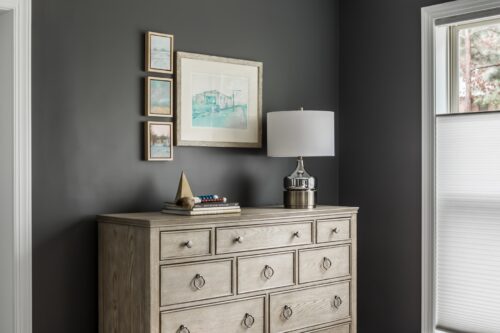 interior design master bedroom light brown wood dresser stack of books framed art collection dark gray walls lamp