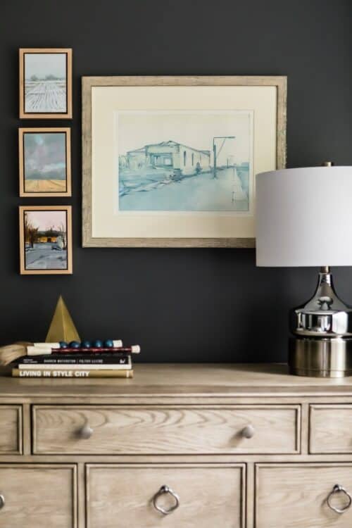interior design master bedroom light brown wood dresser stack of books framed art collection dark gray walls silver lamp